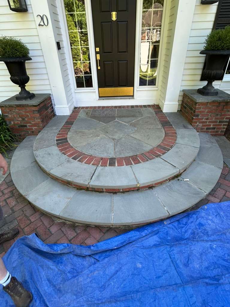 Front porch with curved brick-and-stone steps leading to a dark front door, blue tarp in foreground