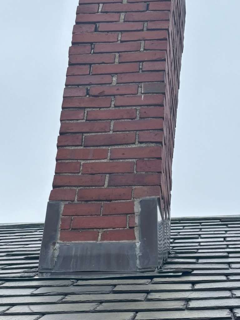 Brick chimney rising from a gray shingled roof against a pale sky