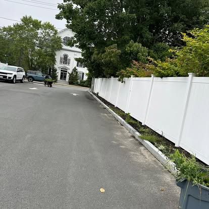 Quiet residential street with parked cars, white fence, and a white house framed by trees.