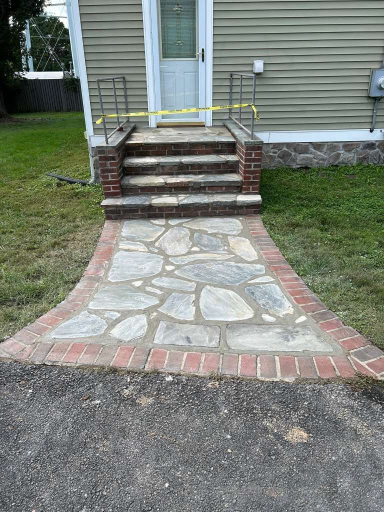 Brick walkway and steps leading to a white front door on a gray house, with a mailbox and handrails.