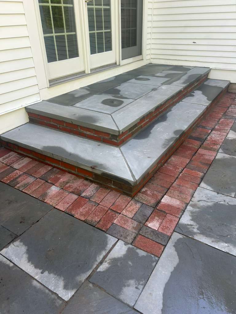 Wet stone patio with brick border and raised steps outside white house siding