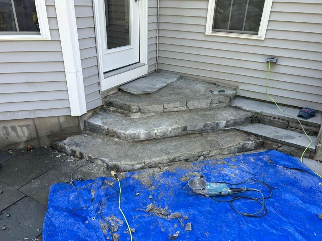 Damaged concrete steps leading to a house door, with a blue tarp covering the ground in front.