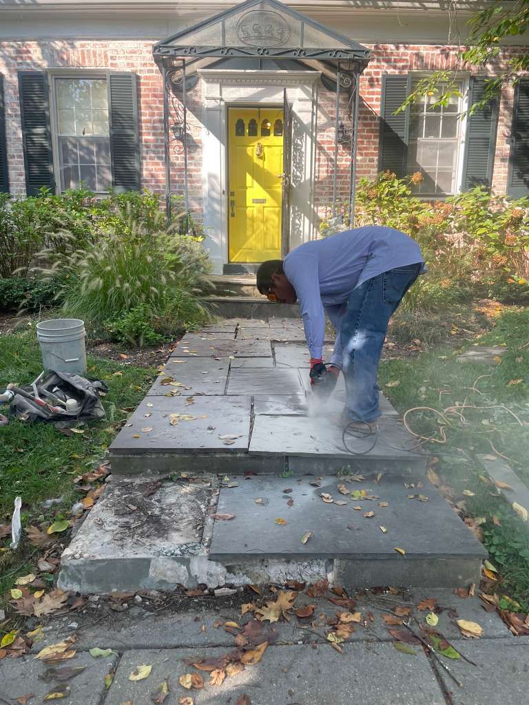 Person laying stone pavers on a front walkway beside a brick house with a yellow door