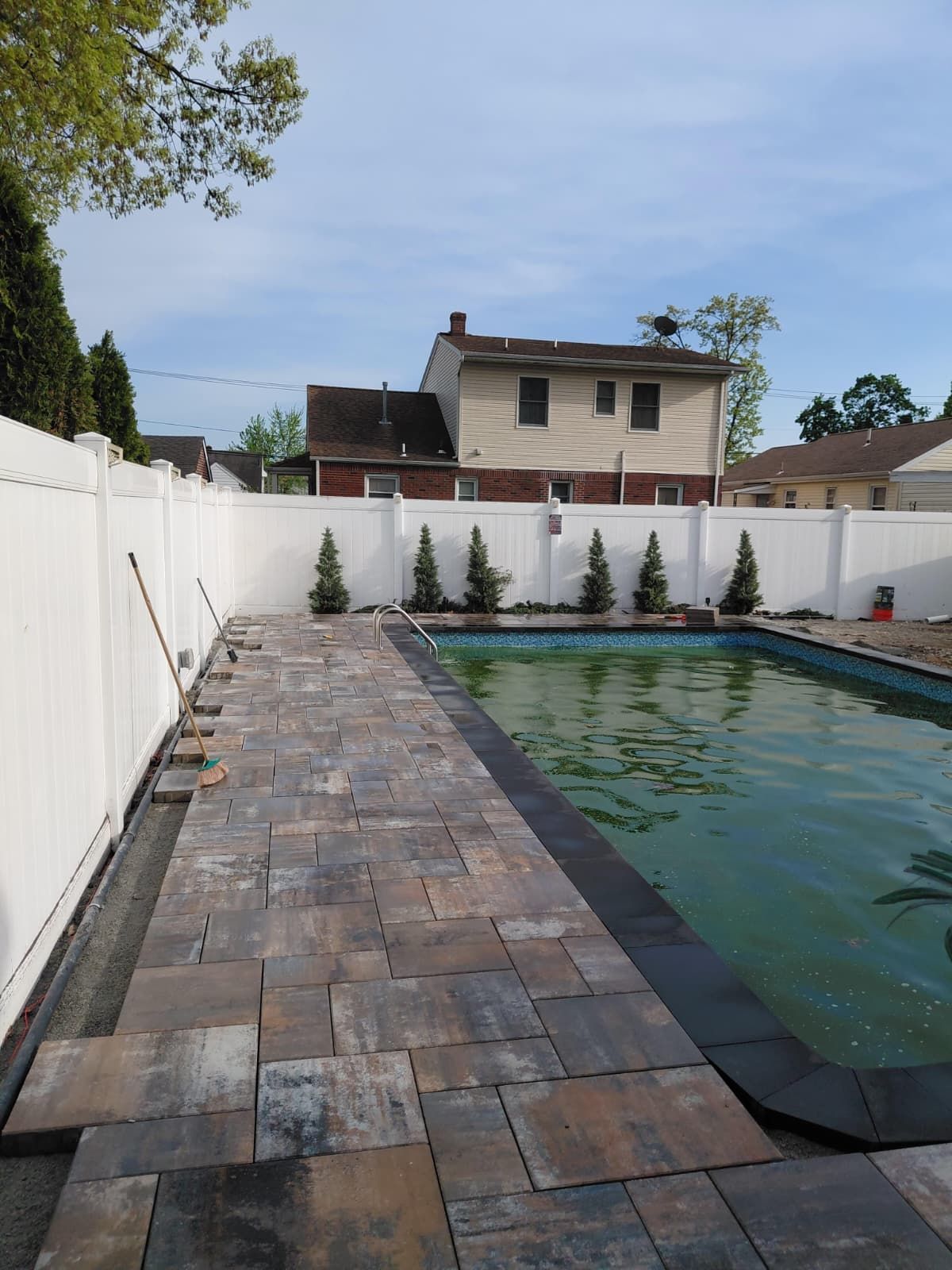 Backyard pool with stone patio, white fence, and small evergreens under a blue sky