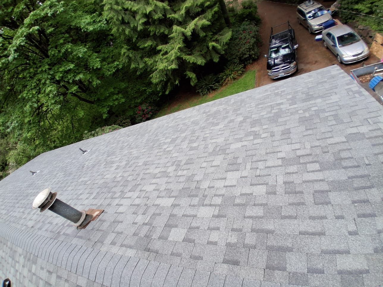 Person standing on a shingled roof near parked cars and trees below