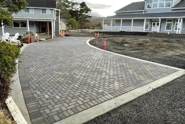 Curved brick driveway between two houses, bordered by concrete and gravel, with orange cones nearby.