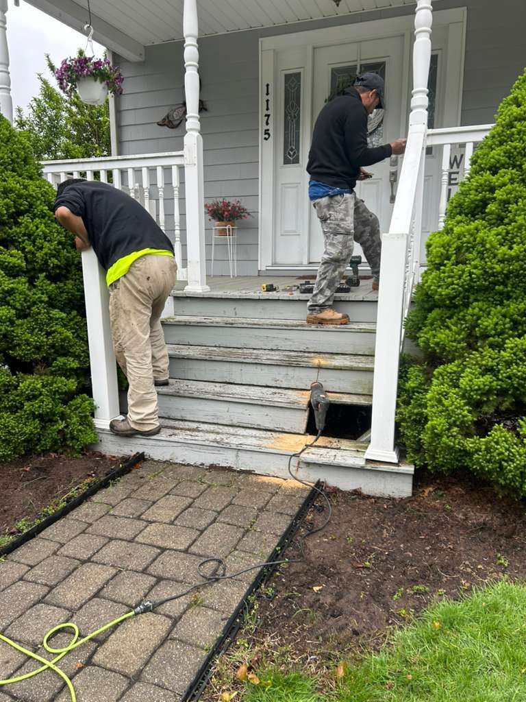 Two workers repair front porch steps and railing on a house.