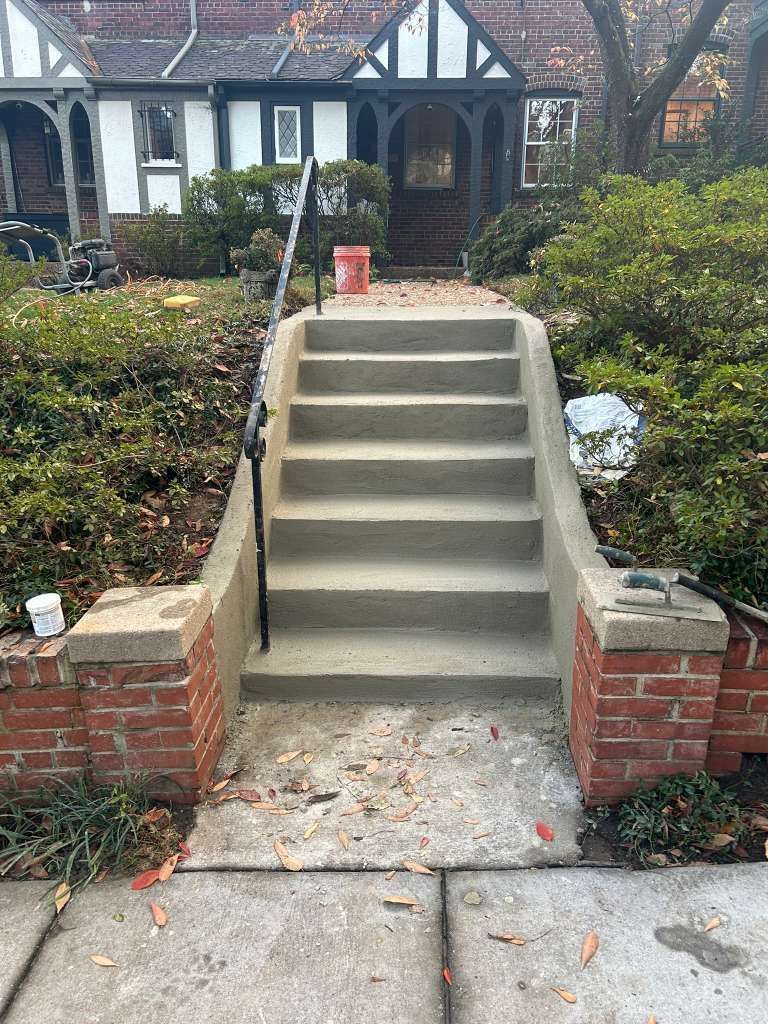 Front walkway with concrete steps leading to a brick house entrance, bordered by shrubs and low brick walls