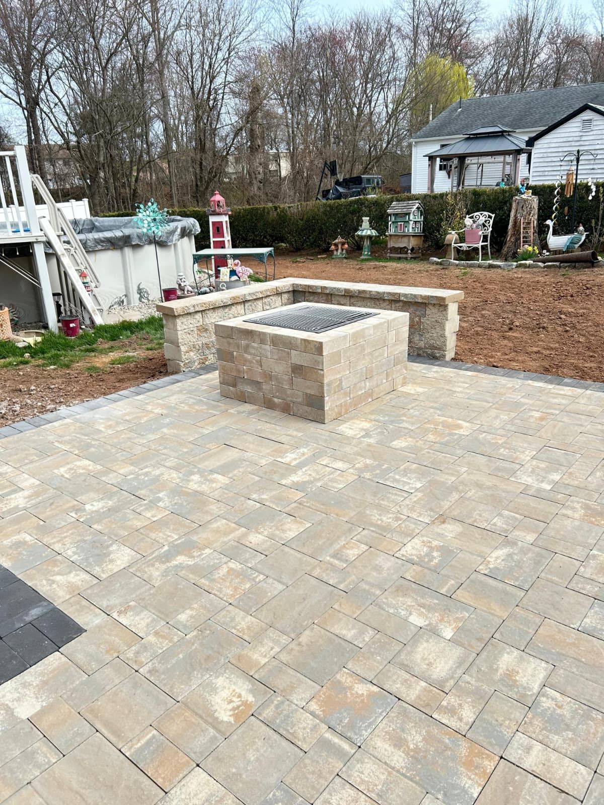 Backyard patio with stone pavers and a raised fire pit, surrounded by trees and a house.