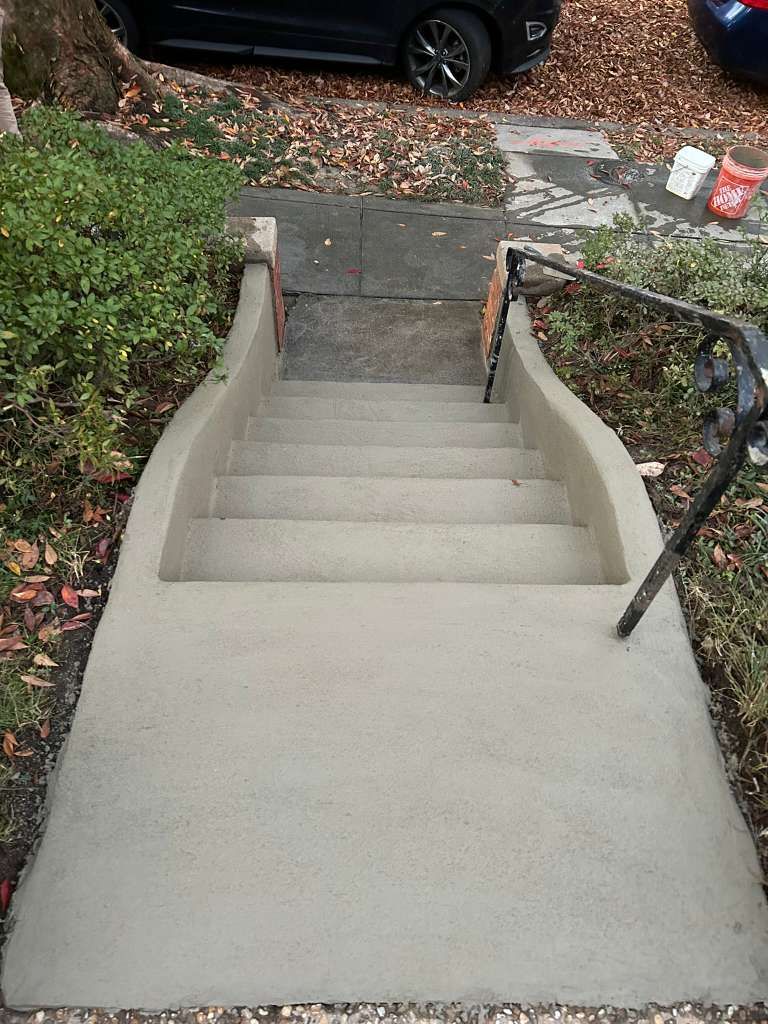 Concrete front steps leading to a sidewalk, with a black handrail and bushes beside them