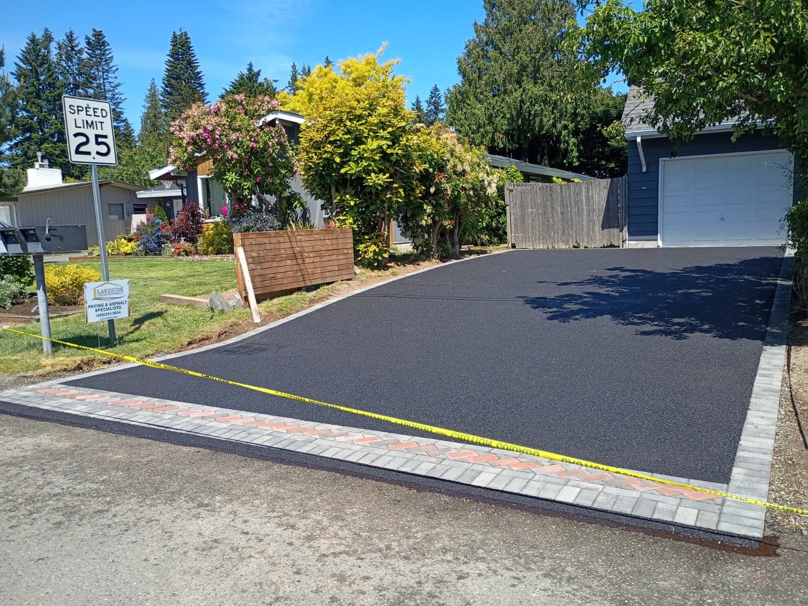 New driveway with black asphalt beside a suburban house, bordered by brick and paver curb stones.