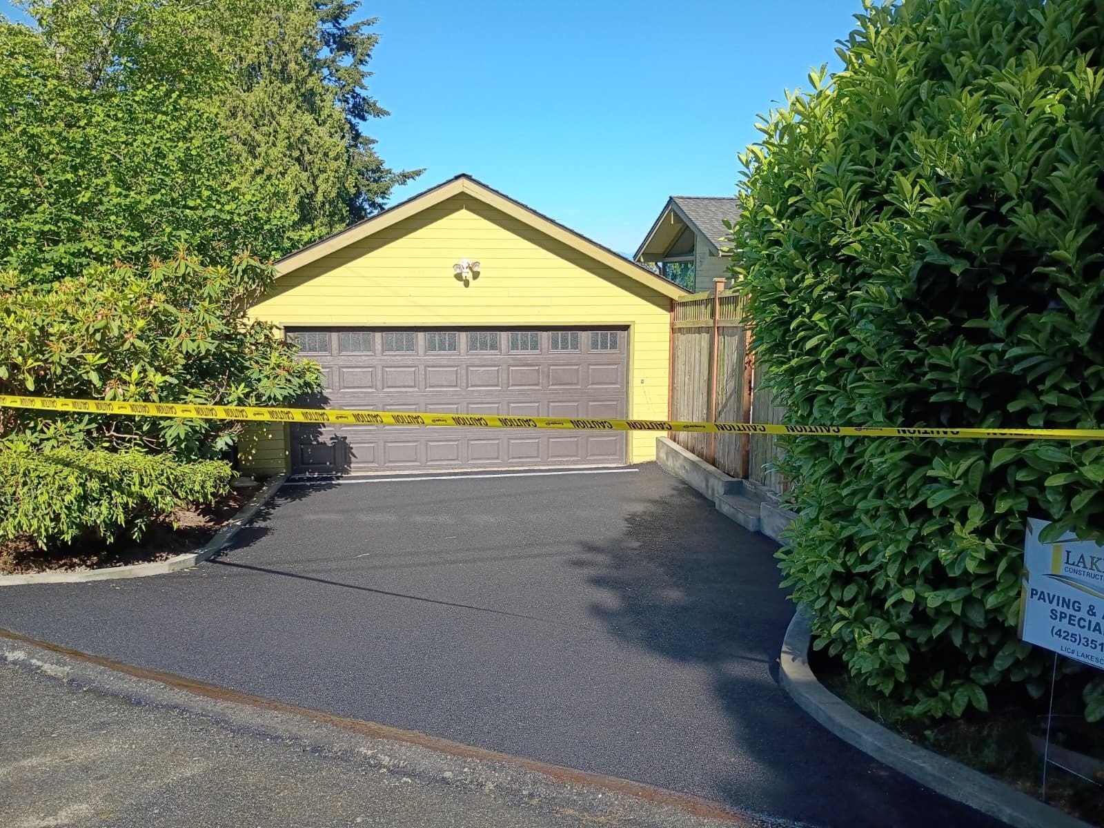 Yellow house with a gray garage door blocked by yellow caution tape, framed by trees and bushes.