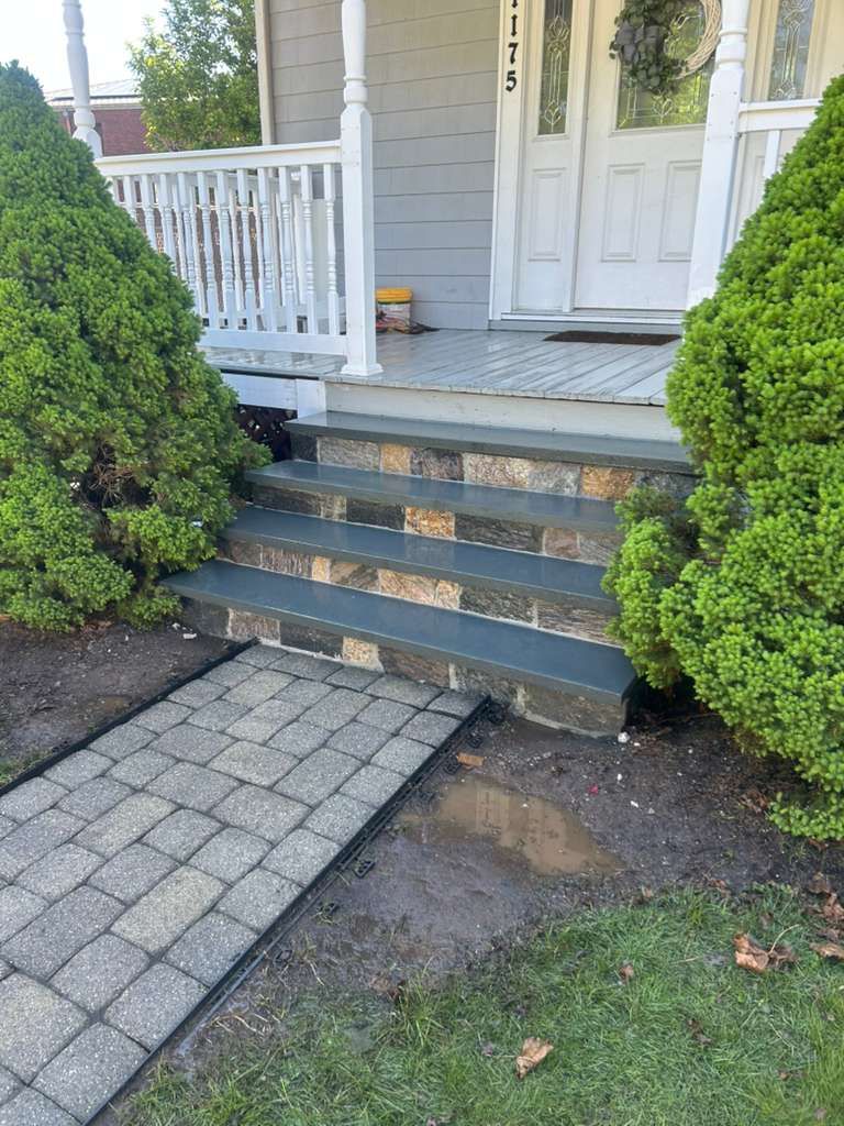 Front steps of a white house with blue trim, flanked by two green shrubs and a stone walkway.