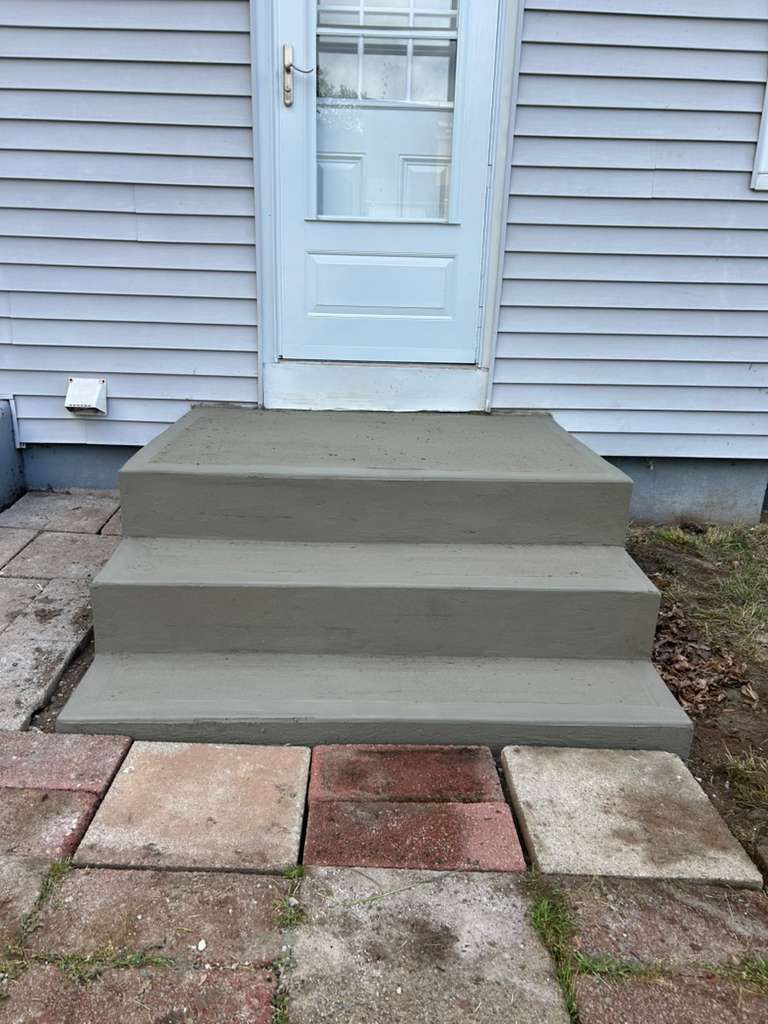 Concrete front steps leading to a white door on a gray-sided house, with stone patio pavers below