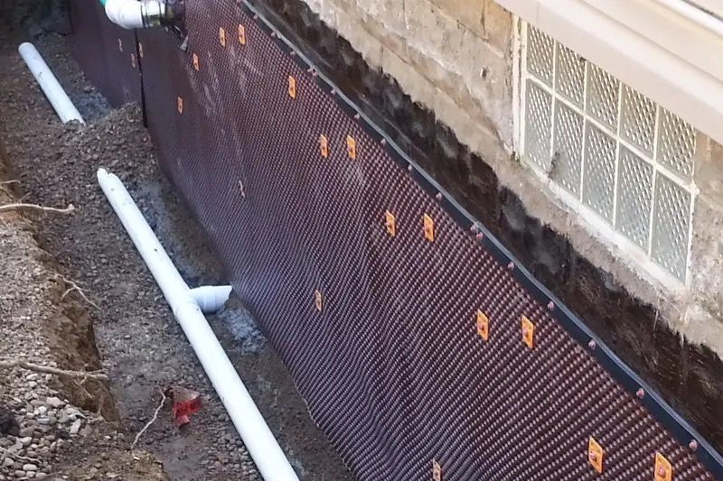 Black drainage mesh along a foundation wall with white pipes and orange clips in a dirt trench