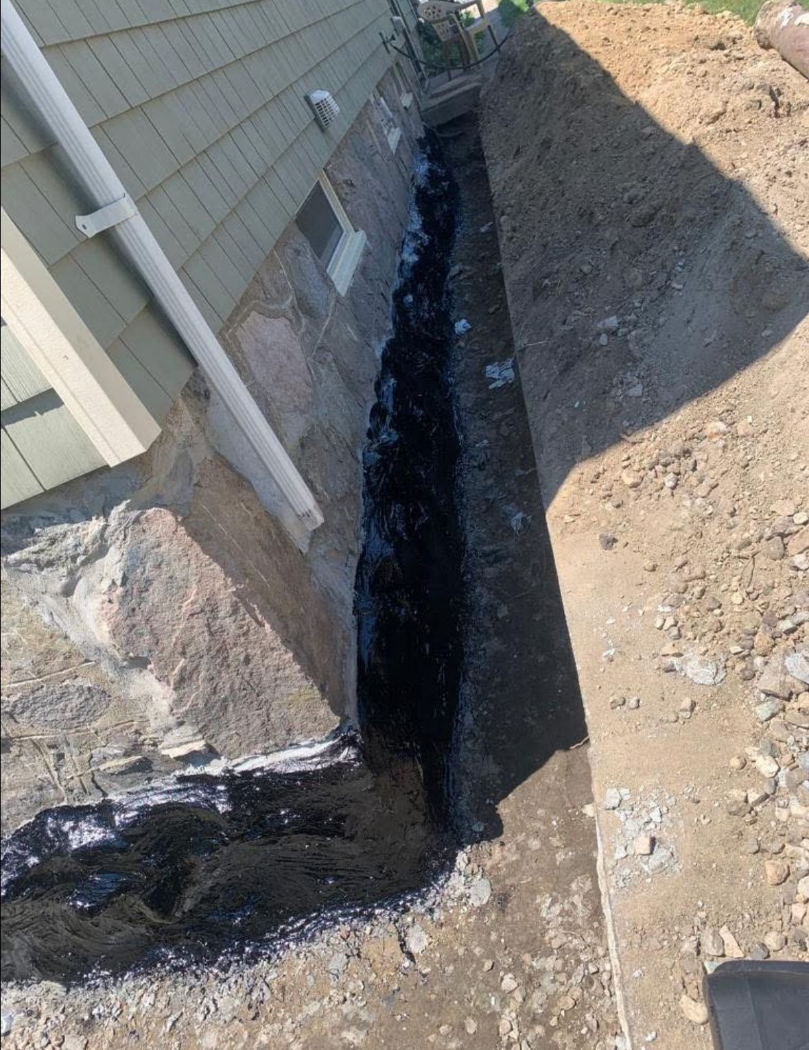 Narrow trench dug beside a house foundation, with black waterproof lining along the wall