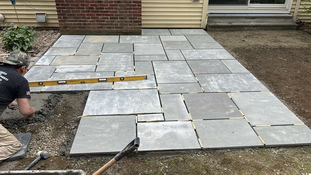 Worker laying gray paver patio beside a house, with tools and leveling bars visible