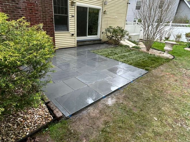 Small backyard patio with large gray tiles beside a house, bordered by grass and shrubs.