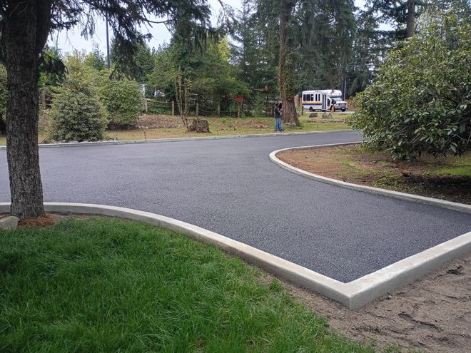 Curved paved driveway in a wooded yard, with grass and shrubs in the foreground and a house in the distance