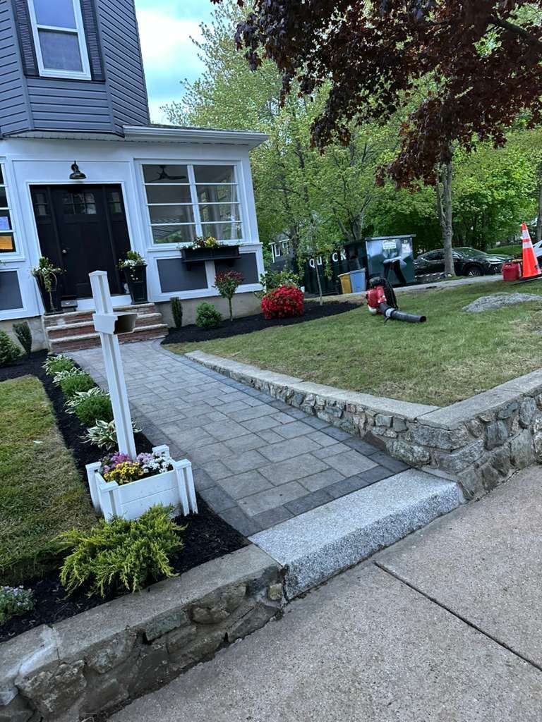 Front yard walkway with pavers, stone edging, flower boxes, and a house porch with red and white flowers