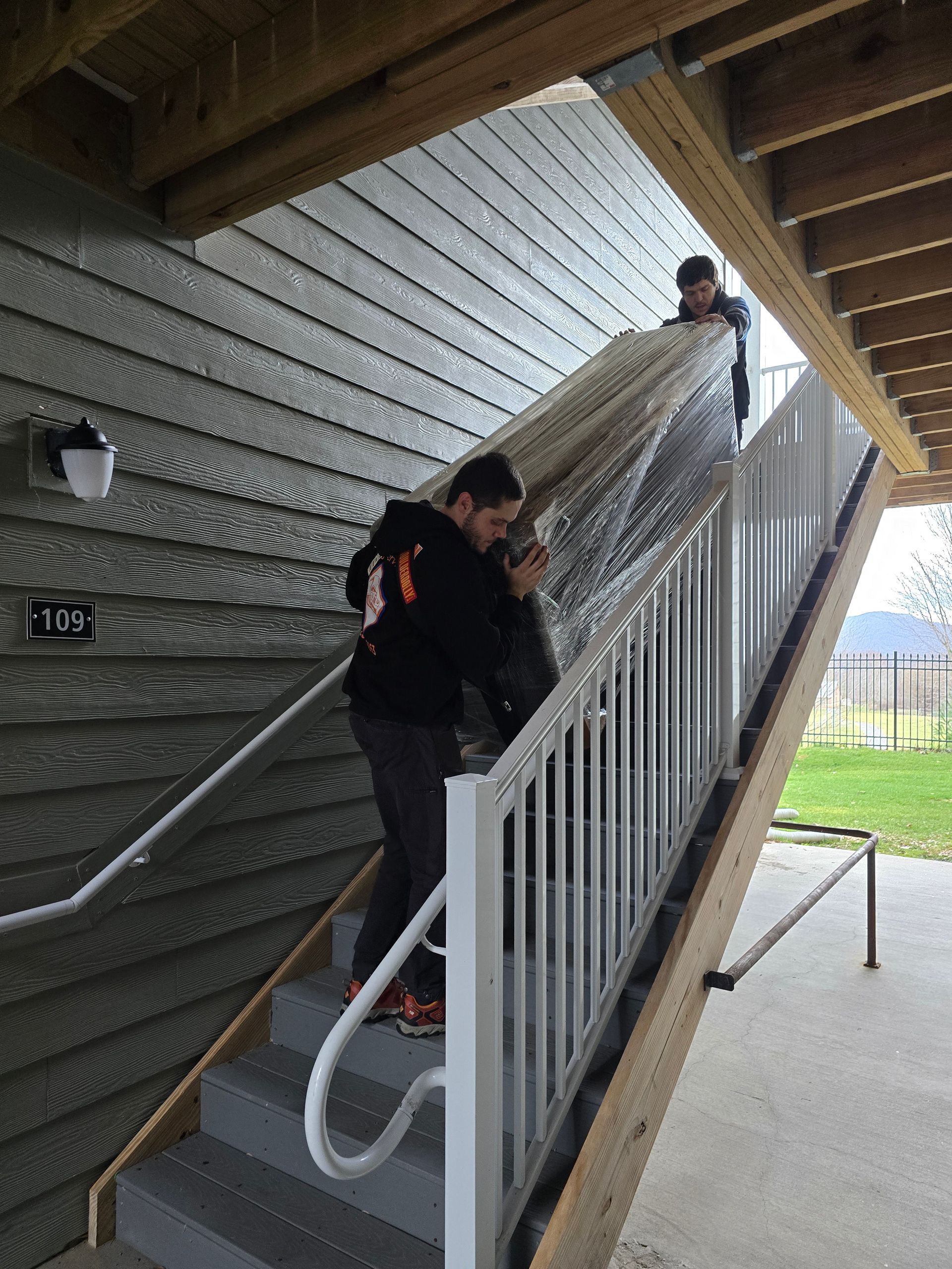 Two people carrying a rolled carpet down outdoor stairs beside a building.