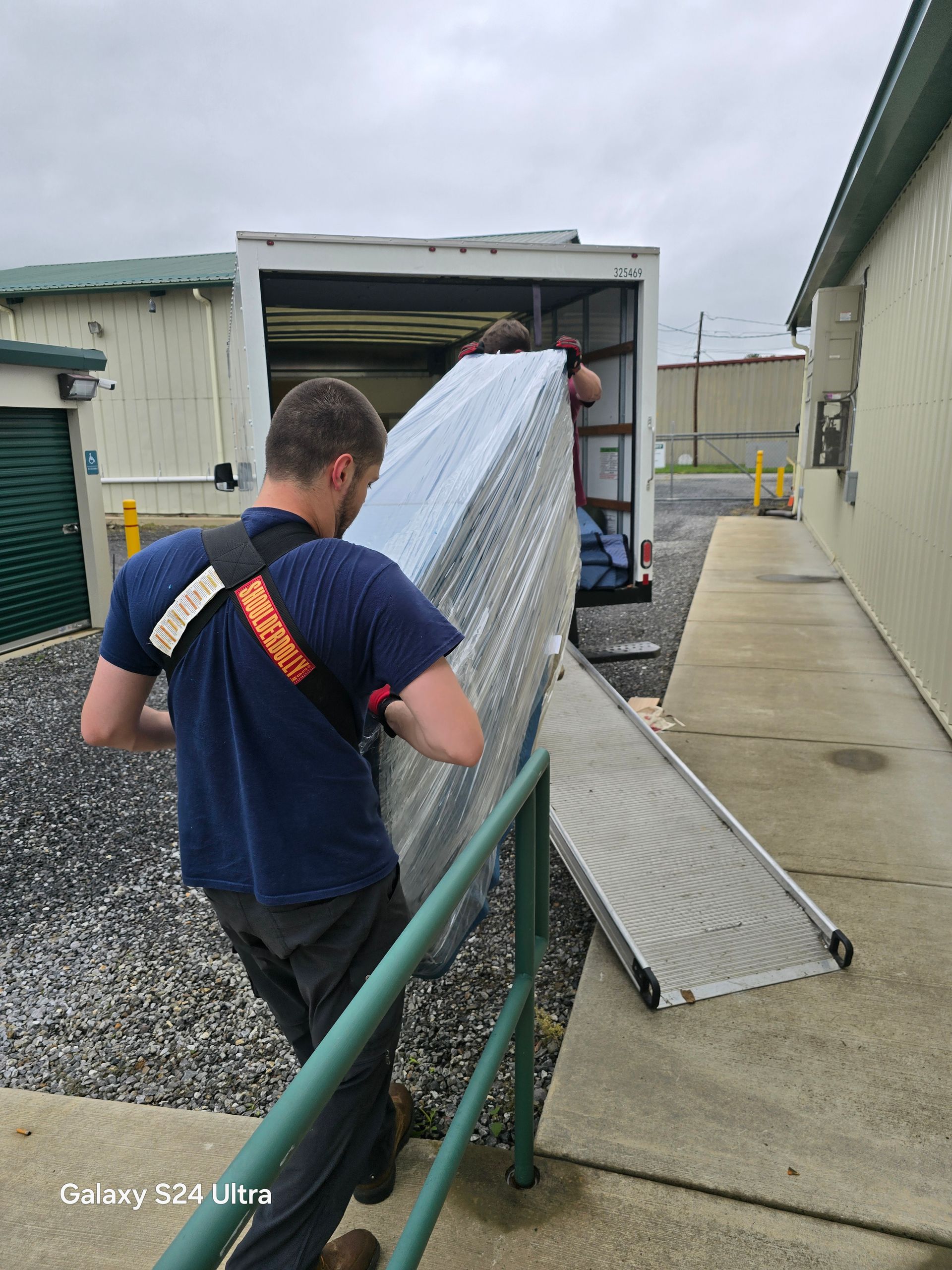Worker unloading a box truck using a metal ramp beside an industrial building