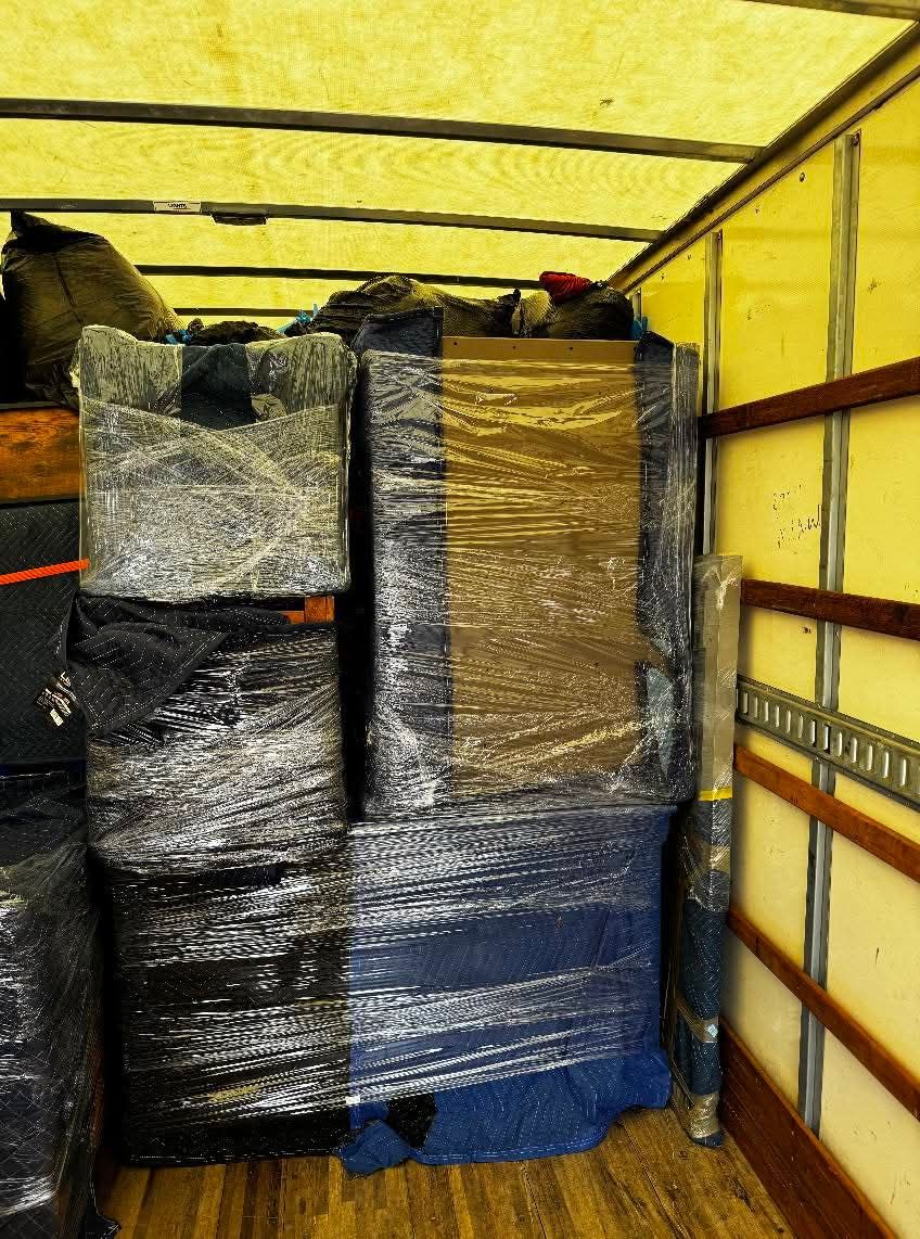 Stacked wrapped cargo in a yellow truck bed, secured with straps and blue padding