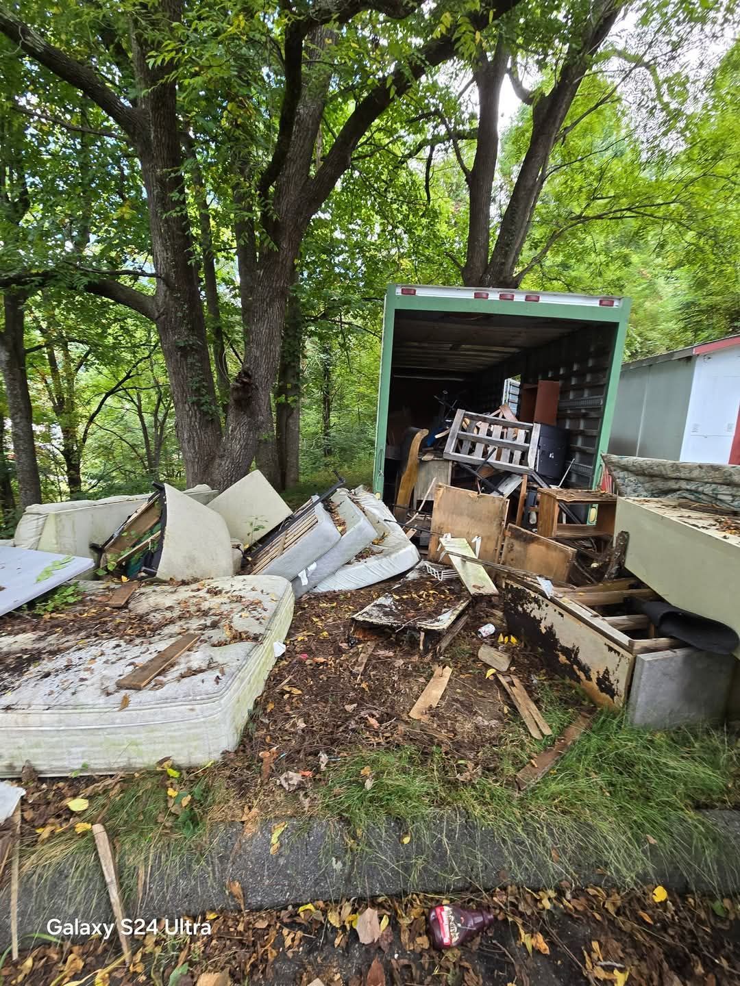 Open green truck and scattered debris on a wooded roadside, with discarded furniture and trash.