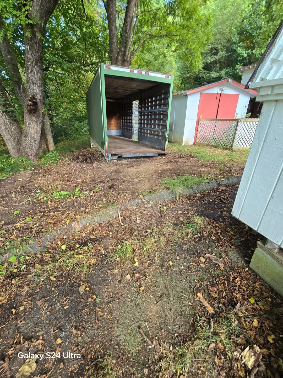 Open shipping container in a wooded yard with muddy ground and nearby sheds.