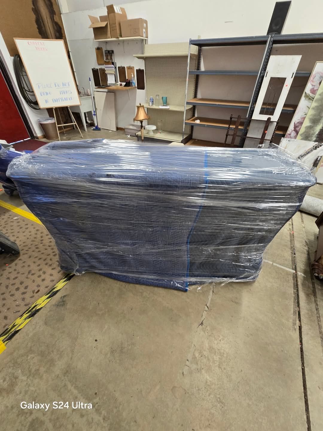 Large blue plastic-wrapped furniture piece in a warehouse, with shelves and boxes in the background