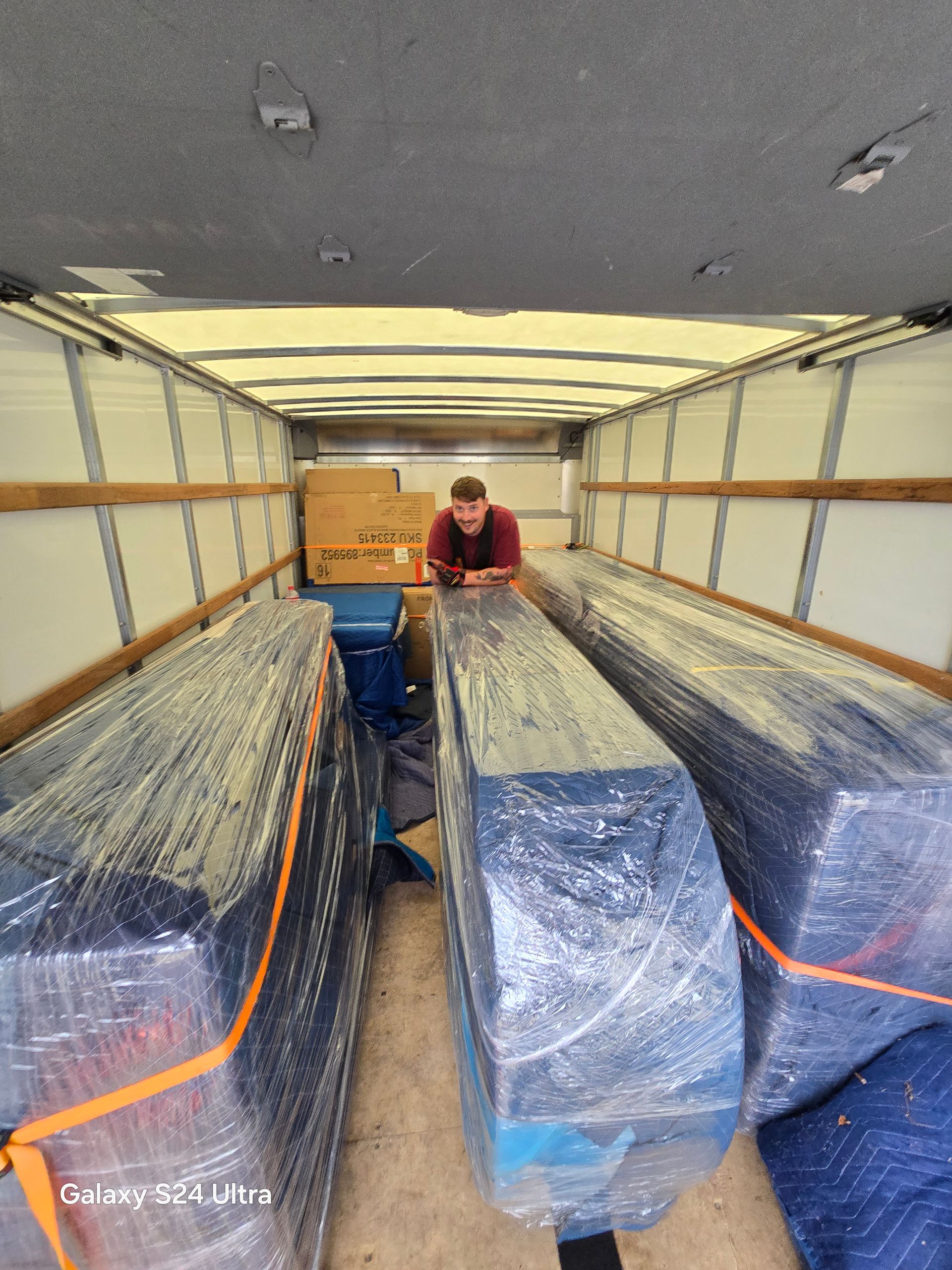 Inside a moving truck, furniture wrapped in plastic and a person securing cargo near the front.