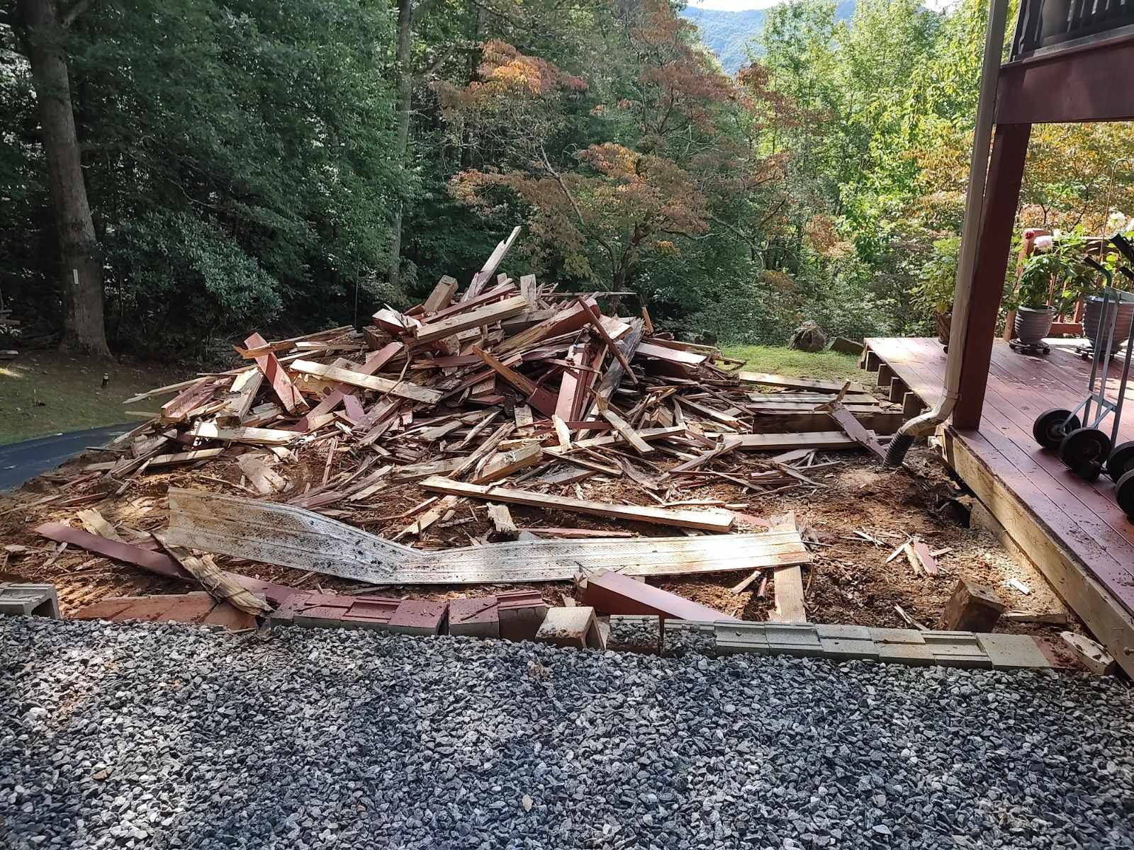 Pile of broken wood and debris in a yard beside a house, with trees in the background.