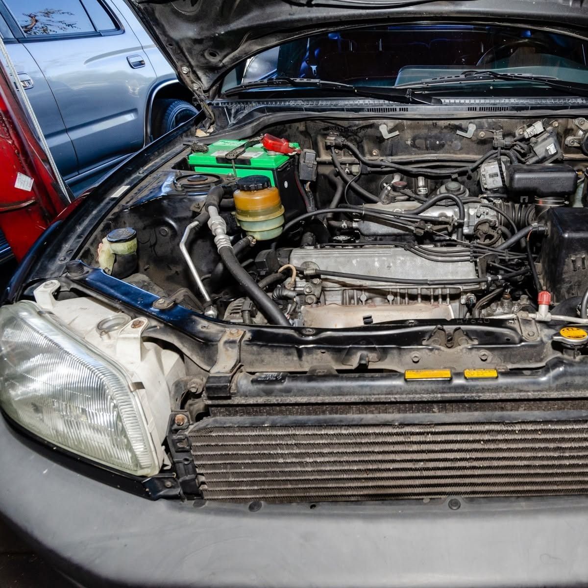 Open car hood showing engine bay with tools and a blue car beside it