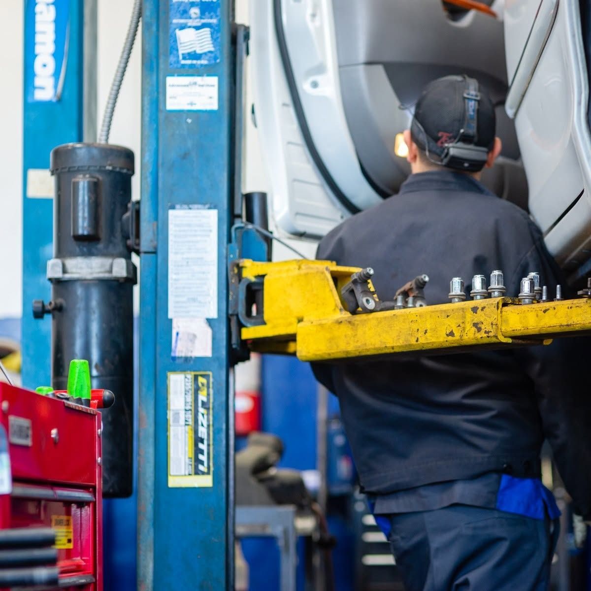 Mechanic operating a car lift in an auto repair shop, with blue machinery and a yellow tool tray nearby