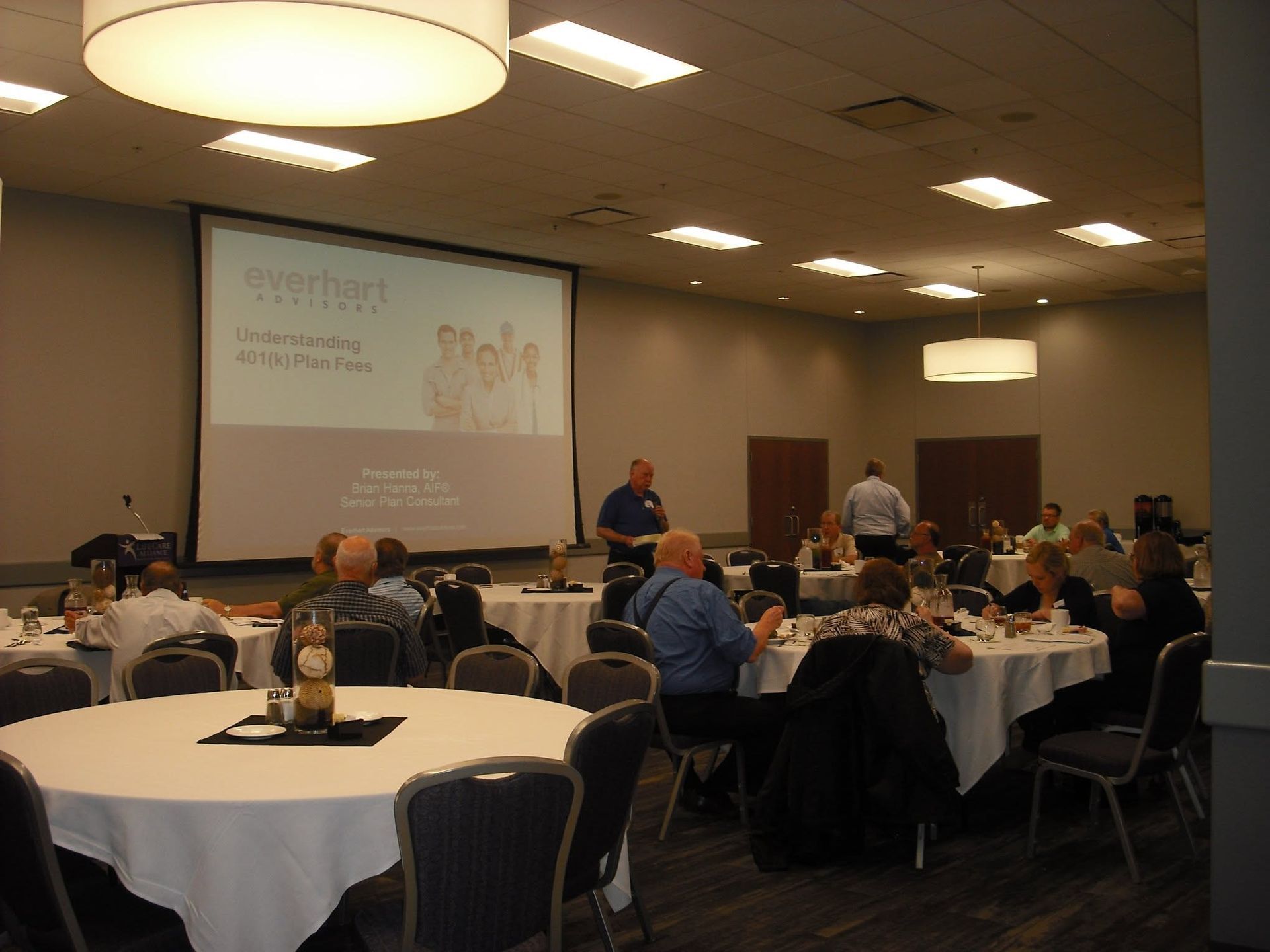 Conference room with round tables, seated attendees, and a presentation projected on a screen