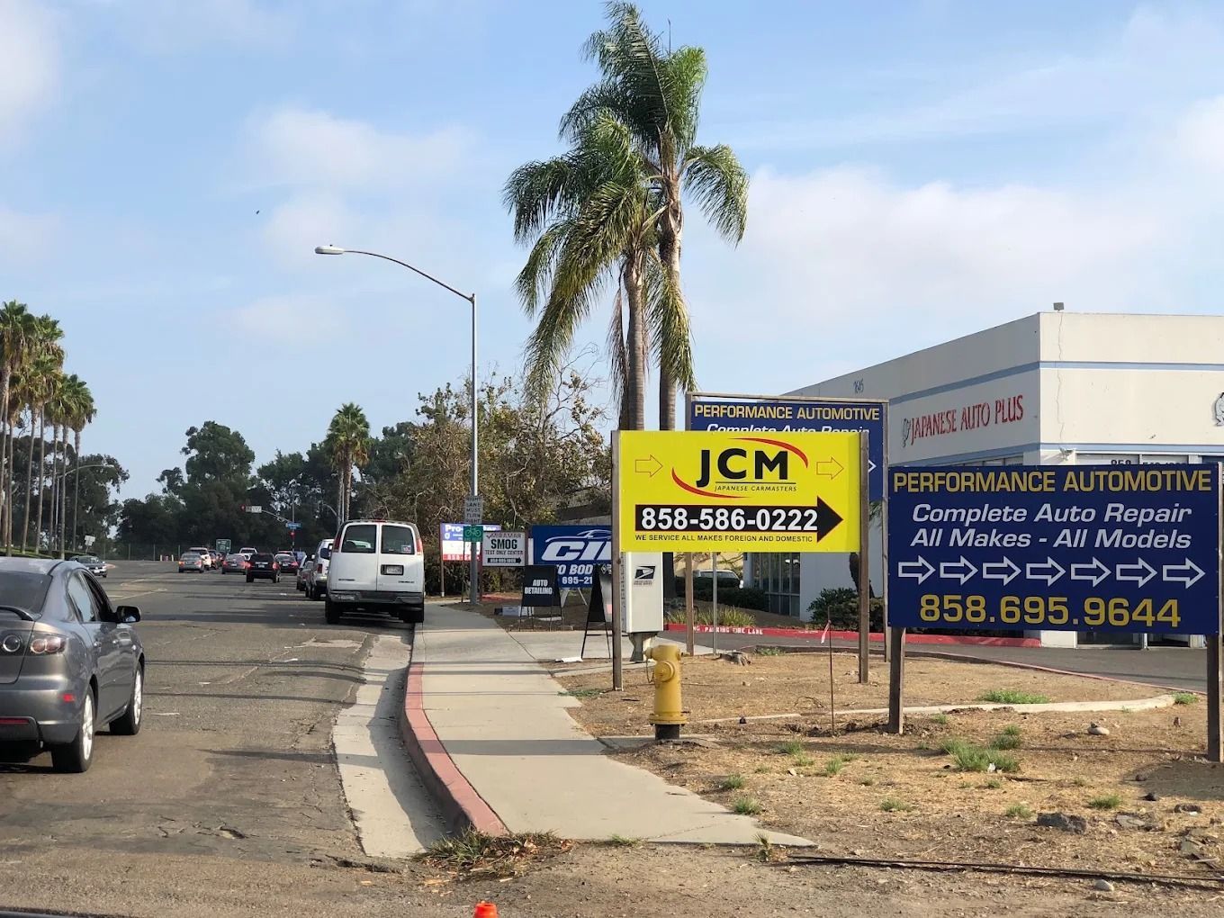 Street with parked cars and real estate signs beside a building, under a partly cloudy sky