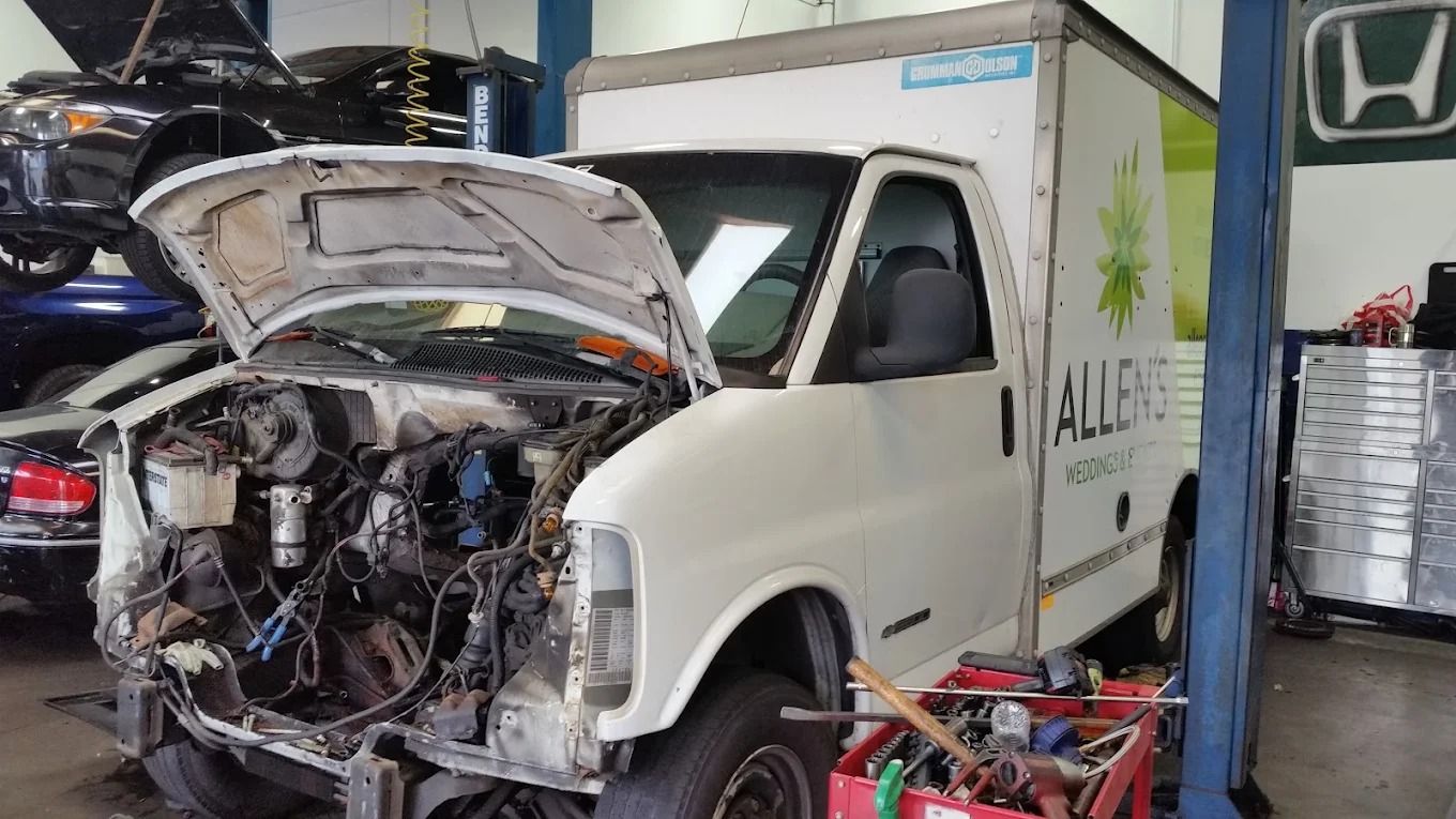 White van with open hood in a repair garage, front end exposed and damaged, surrounded by tools and equipment