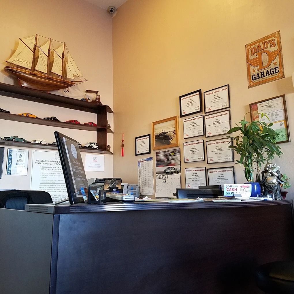Small office reception desk with certificates, a model ship, and potted plant on beige walls.