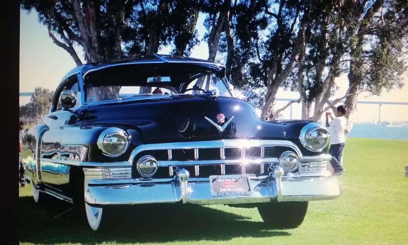 Classic black vintage car parked on grass under trees, front view with chrome details and blue sky.