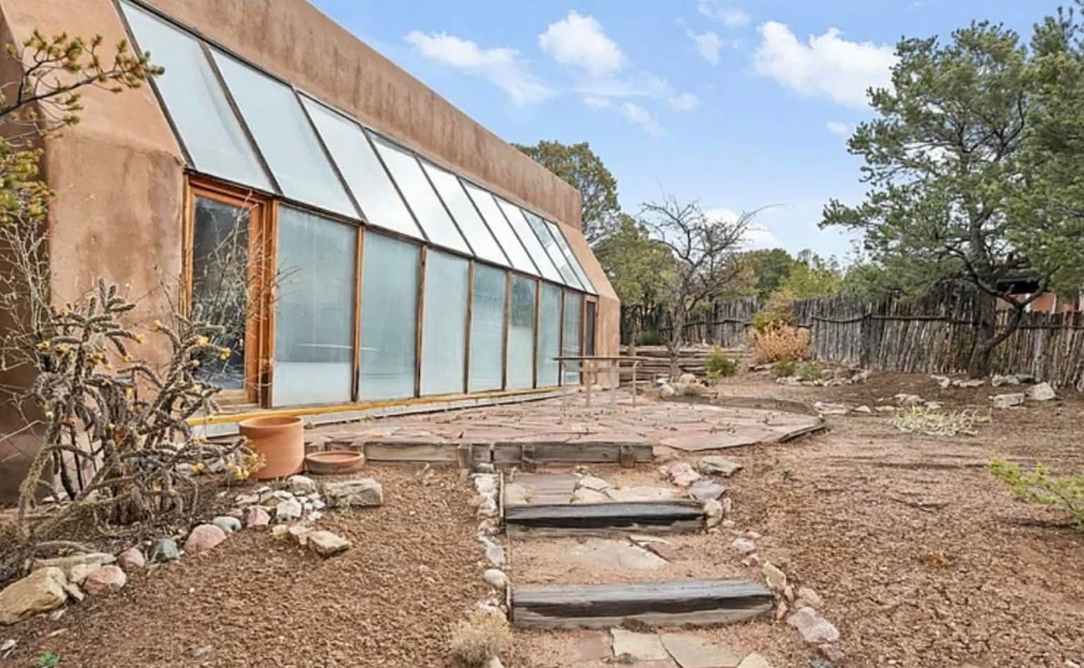 Rustic greenhouse with slanted glass panels beside a stone path in a dry, wooded yard.
