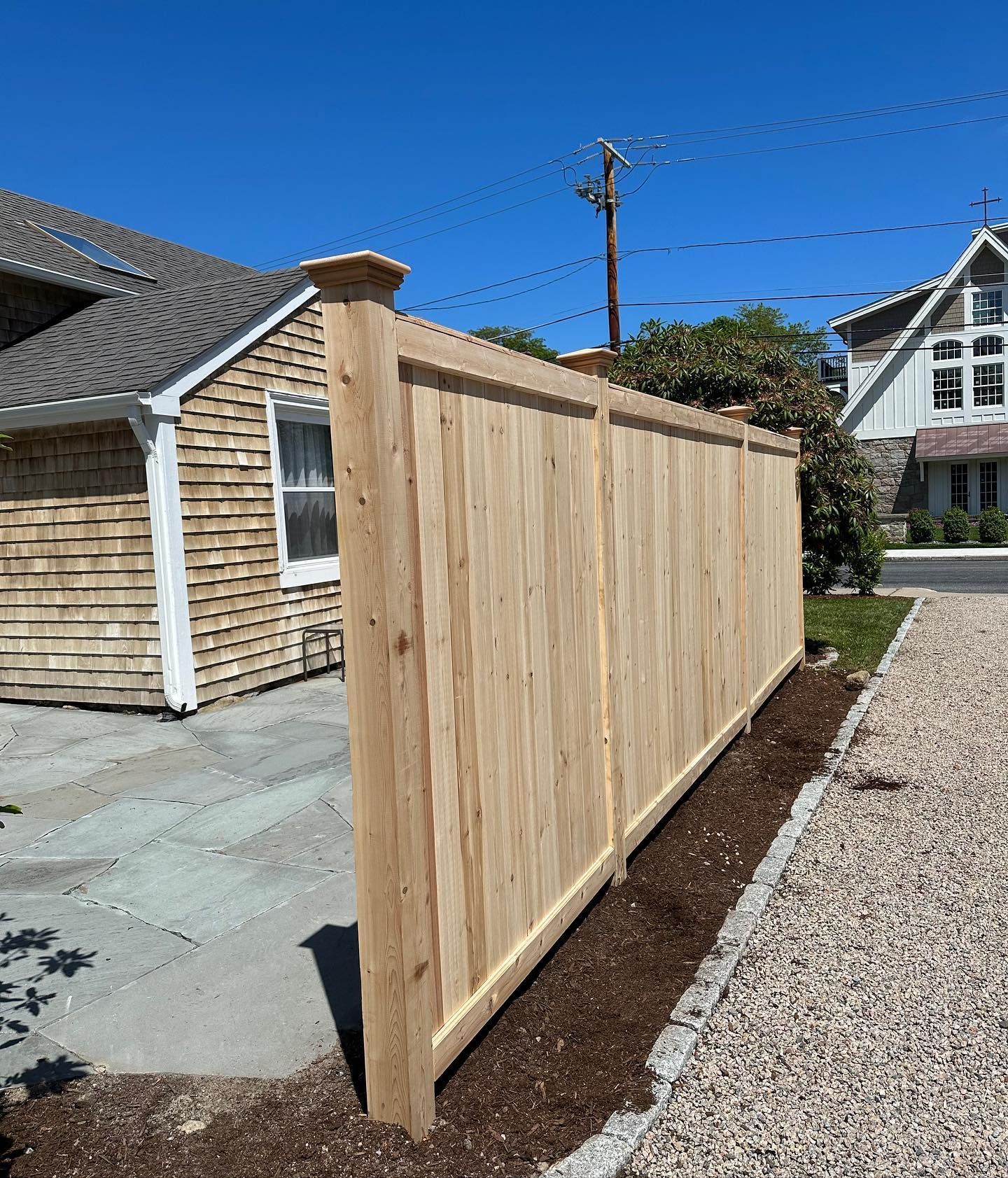 A wooden fence is sitting next to a gravel driveway in front of a house.