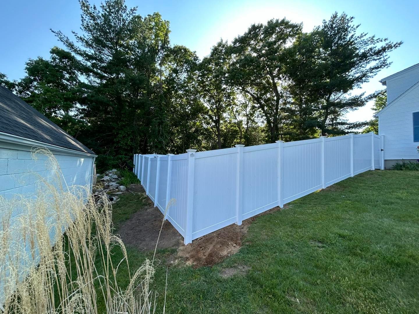 A white vinyl fence is surrounded by grass and trees in a backyard.