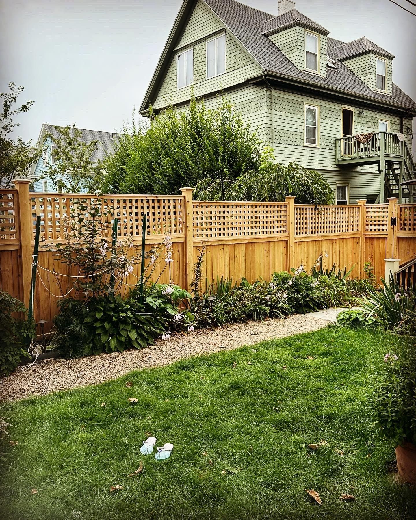 Lush green backyard with a wooden fence, a garden path, and a light-green house in the background.