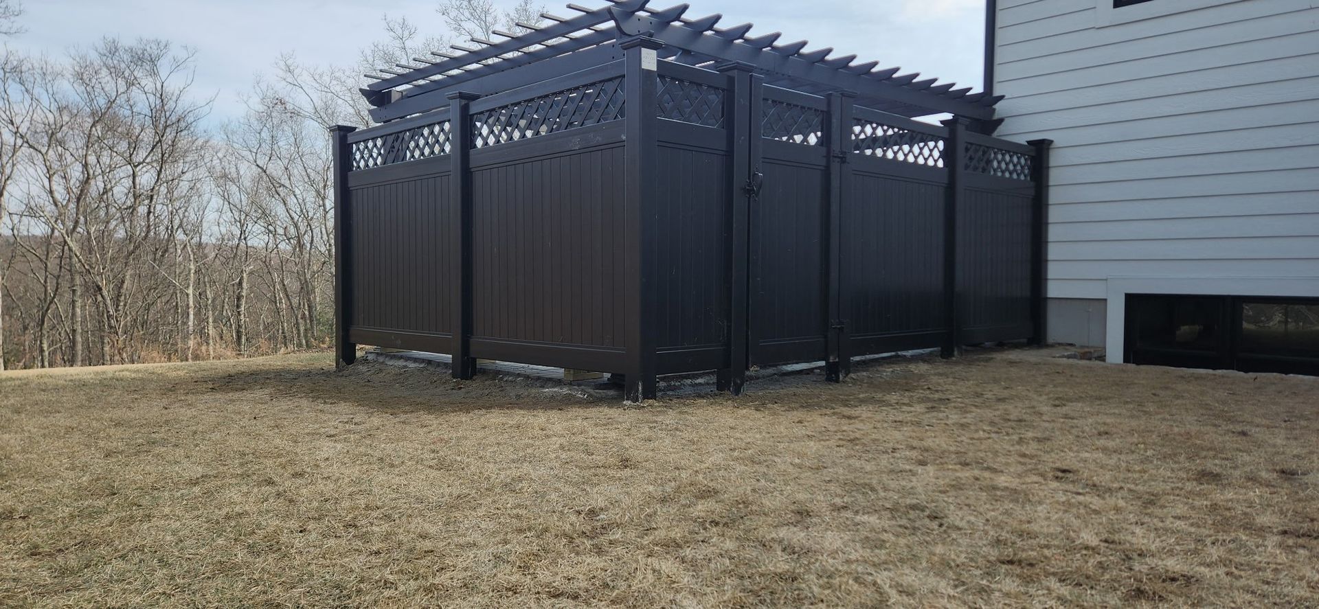 A black fenced enclosure with a pergola roof in a yard, next to a white house.