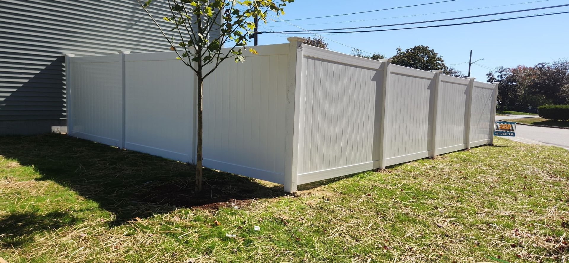 White fence bordering a grassy area next to a building and street with a tree in the foreground.