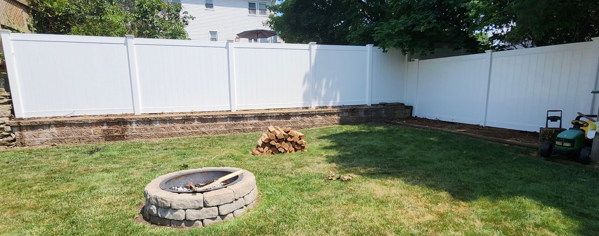Backyard with white fence, grass lawn, and a circular stone fire pit in the foreground