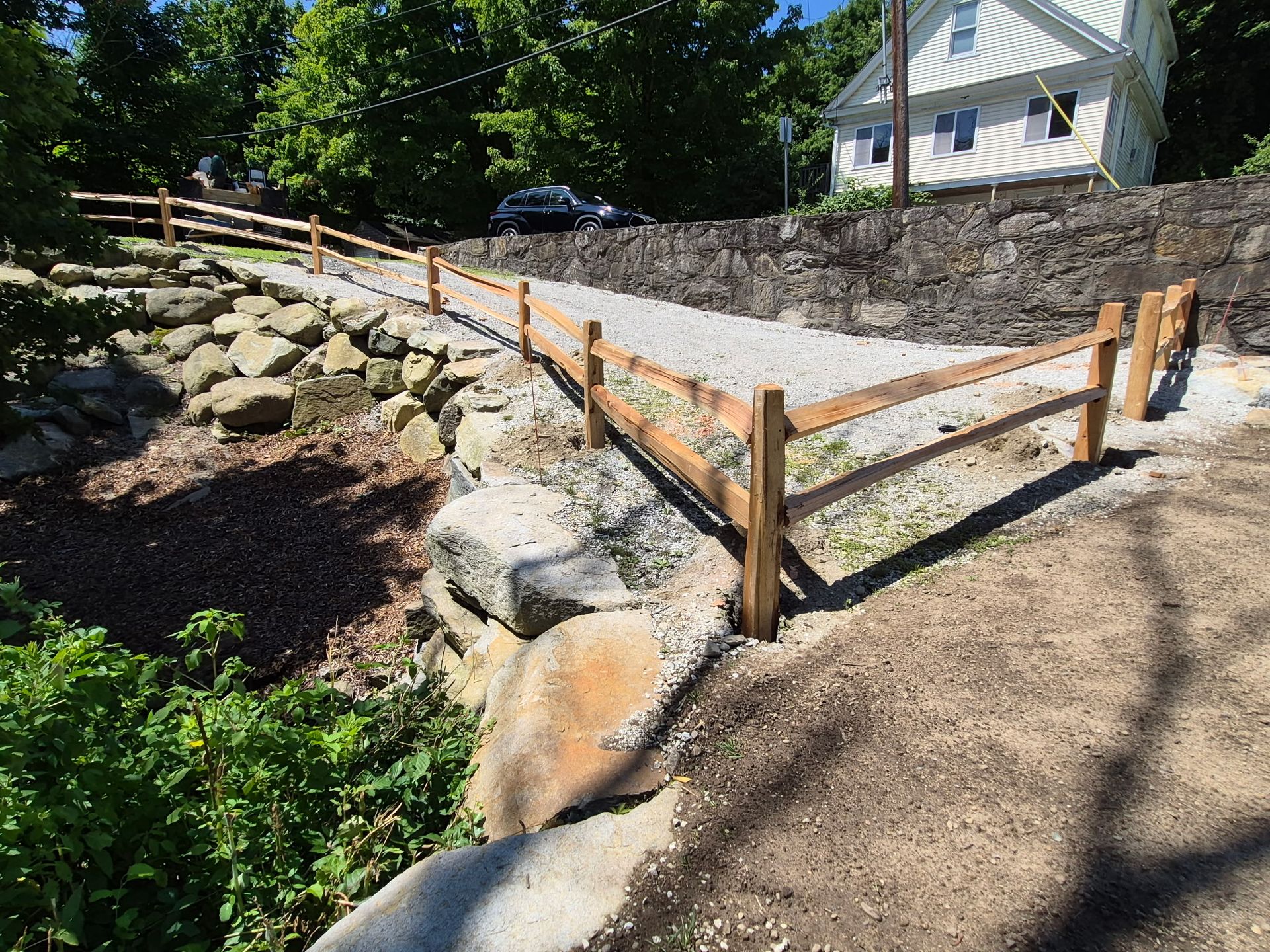 Wooden rail fence bordering a sloped stone retaining wall in a sunny yard with a house in the background