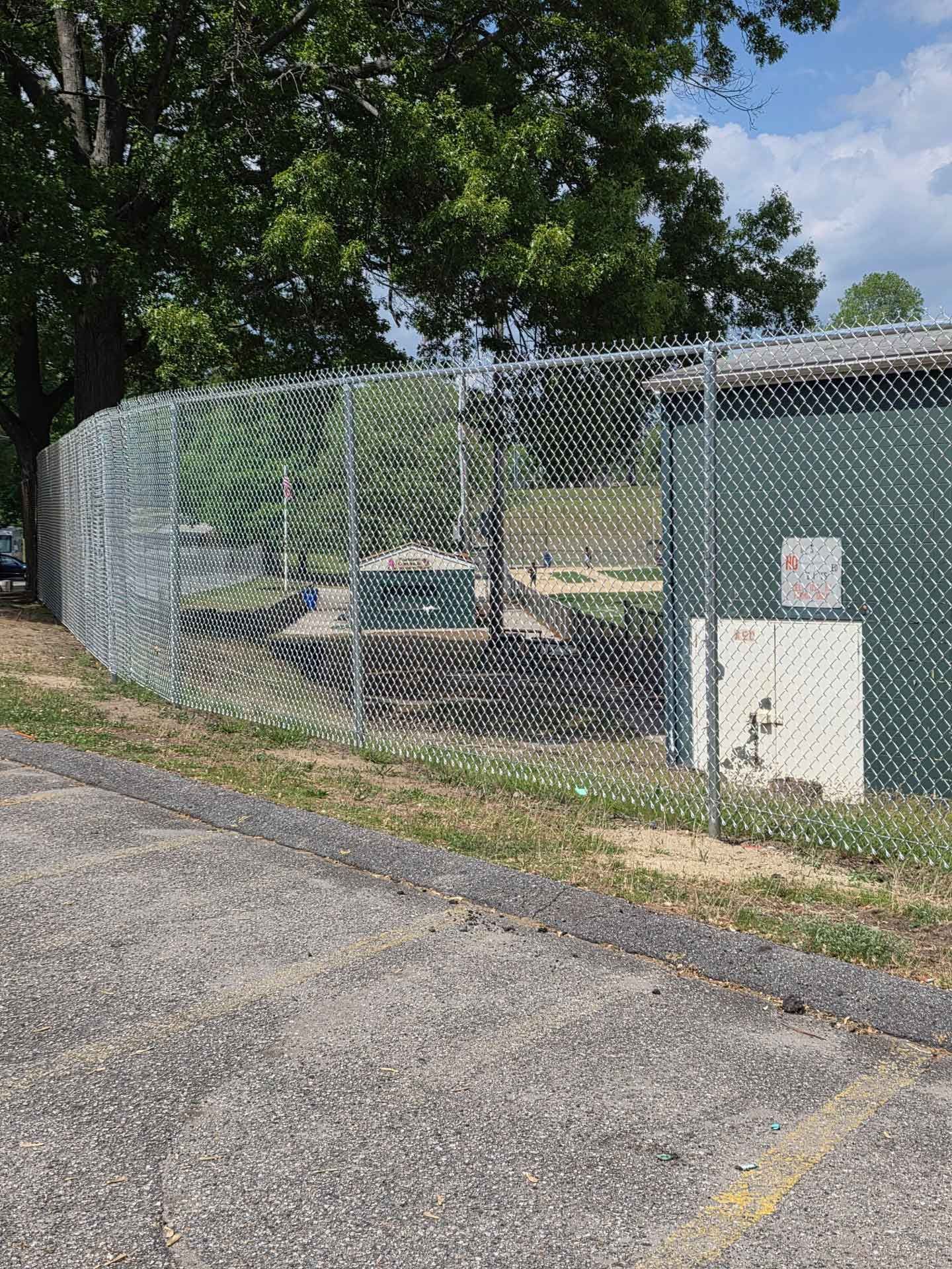 Chain-link fenced utility area beside a parking lot under trees on a sunny day