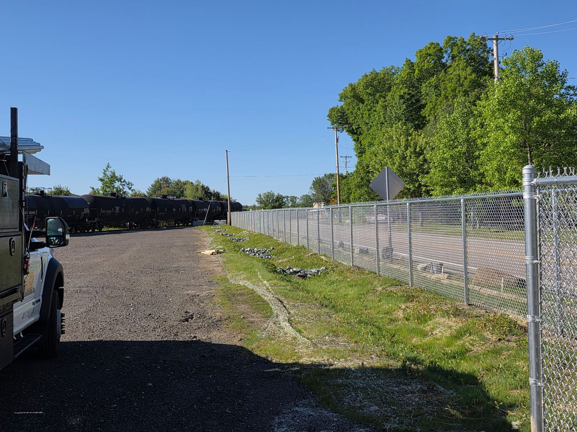 Gravel lot beside a chain-link fence and green trees under a clear blue sky