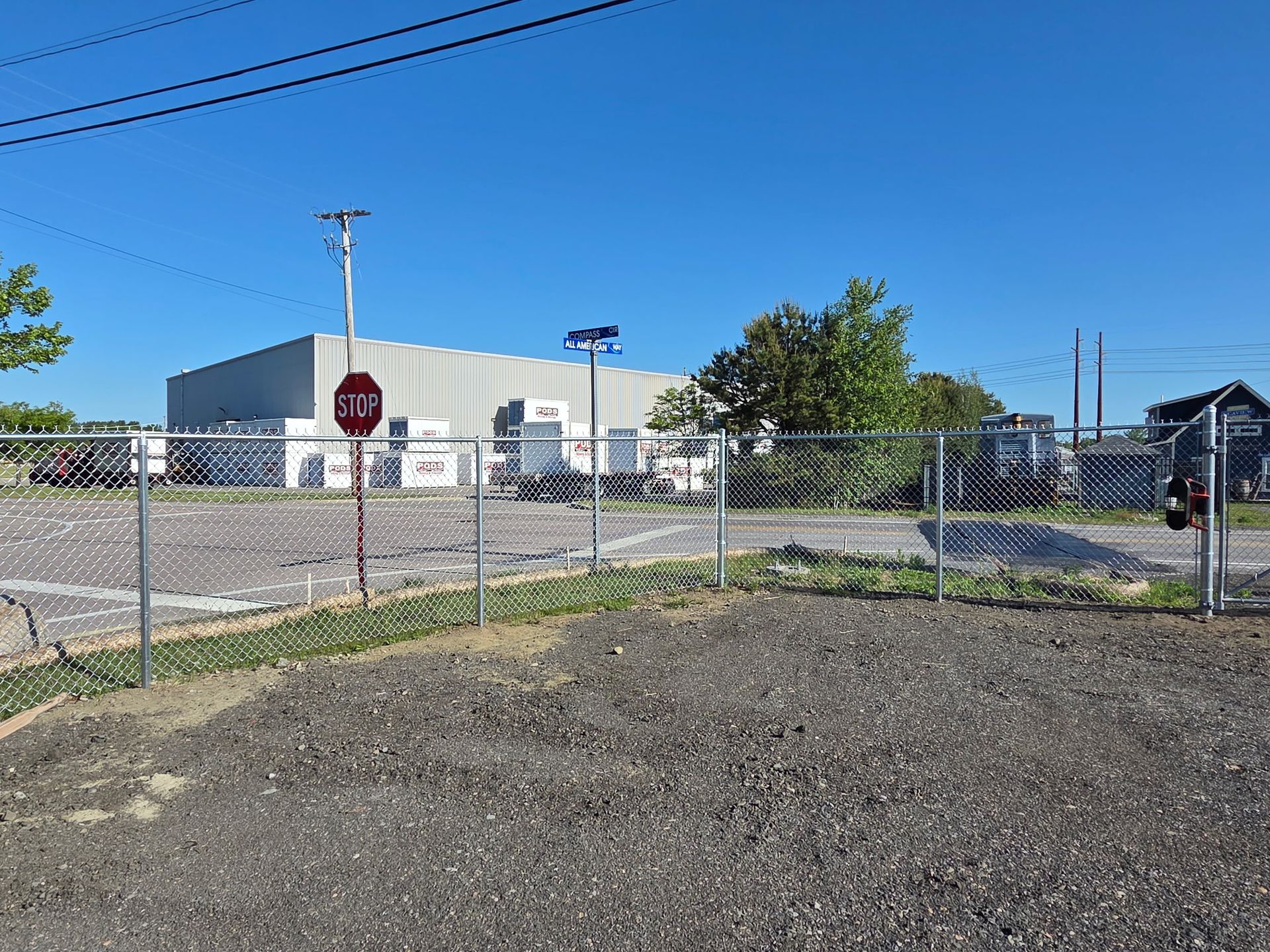 Chain-link fence beside a gravel lot, with a warehouse building and stop sign under a clear blue sky.